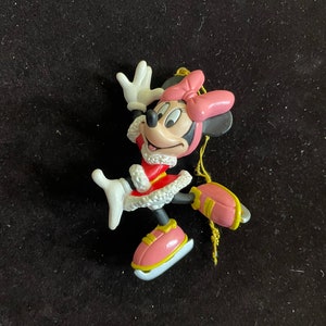May include: A pink and white plastic Christmas ornament featuring Minnie Mouse ice skating. She is wearing a red and white dress with a white fur trim and a pink bow.