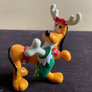 Può includere: Una statuetta in ceramica di Pluto vestito da renna. Indossa un cappello da Babbo Natale rosso e bianco, una sciarpa verde e bianca e un collare rosso e bianco. Tiene un osso bianco nella zampa destra.