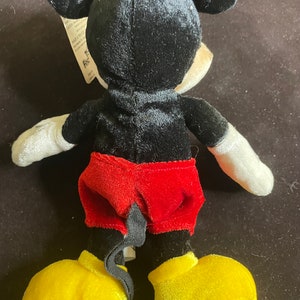 K&ouml;nnte beinhalten: Ein schwarzes und rotes Pl&uuml;sch-Mickey-Mouse-Spielzeug mit gelben Schuhen. Das Spielzeug steht auf einer braunen Oberfl&auml;che.