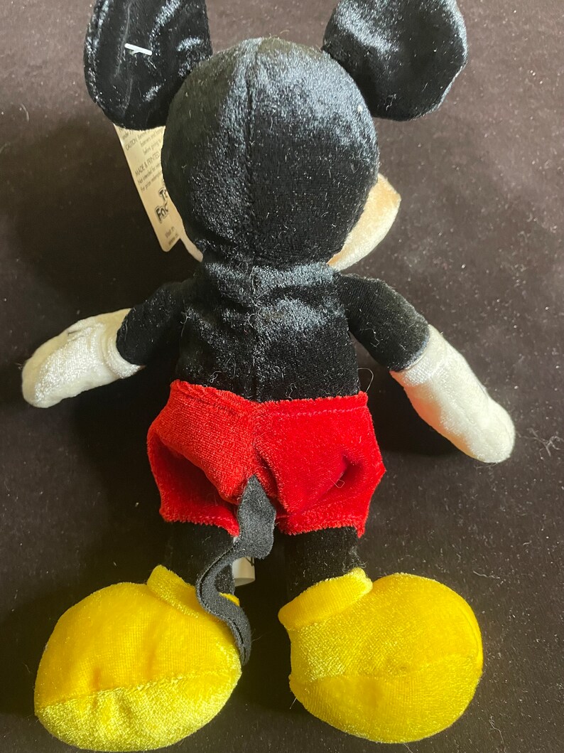 K&ouml;nnte beinhalten: Ein schwarzes und rotes Pl&uuml;sch-Mickey-Mouse-Spielzeug mit gelben Schuhen. Das Spielzeug steht auf einer braunen Oberfl&auml;che.