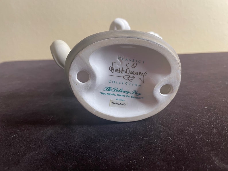 May include: White ceramic figurine of Mickey Mouse dressed as a delivery boy. The base of the figurine is white with a green label that reads "CLASSICS Walt Disney COLLECTION The Delivery Boy "Hey Minnie, Wanna Go Steppin'" &copy; Disney THAILAND"