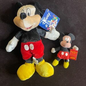 K&ouml;nnte beinhalten: Zwei Pl&uuml;sch-Mickey-Mouse-Spielzeuge. Der gr&ouml;&szlig;ere Mickey tr&auml;gt eine rote Hose mit wei&szlig;en Kreisen und gelben Schuhen. Der kleinere Mickey tr&auml;gt eine rote Hose mit wei&szlig;en Kreisen und gelben Schuhen. Beide Mickey-Mouse-Spielzeuge haben schwarzes Fell und gro&szlig;e Ohren.
