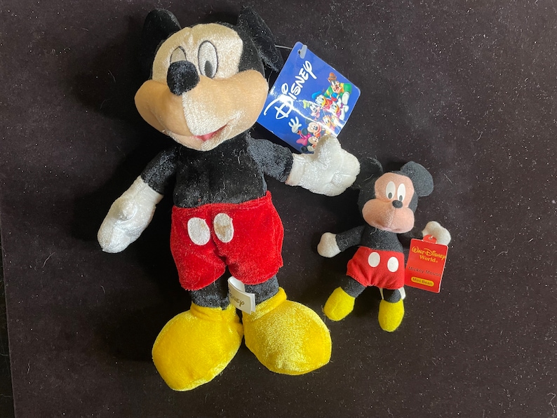 K&ouml;nnte beinhalten: Zwei Pl&uuml;sch-Mickey-Mouse-Spielzeuge. Der gr&ouml;&szlig;ere Mickey tr&auml;gt eine rote Hose mit wei&szlig;en Kreisen und gelben Schuhen. Der kleinere Mickey tr&auml;gt eine rote Hose mit wei&szlig;en Kreisen und gelben Schuhen. Beide Mickey-Mouse-Spielzeuge haben schwarzes Fell und gro&szlig;e Ohren.