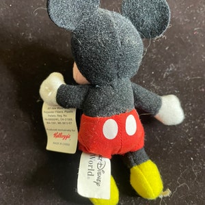 K&ouml;nnte beinhalten: Ein schwarzes und rotes Pl&uuml;sch-Mickey-Mouse-Spielzeug mit gelben Schuhen. Das Spielzeug tr&auml;gt ein rot-wei&szlig;es Hemd mit zwei wei&szlig;en Kreisen. Das Spielzeug hat ein Etikett mit der Aufschrift "Disney World" und "Kellogg's".