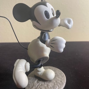 May include: A black and white ceramic figurine of Mickey Mouse running. The figurine is in a classic cartoon style and is posed with his right arm raised and his left leg forward. The figurine is standing on a gray base.