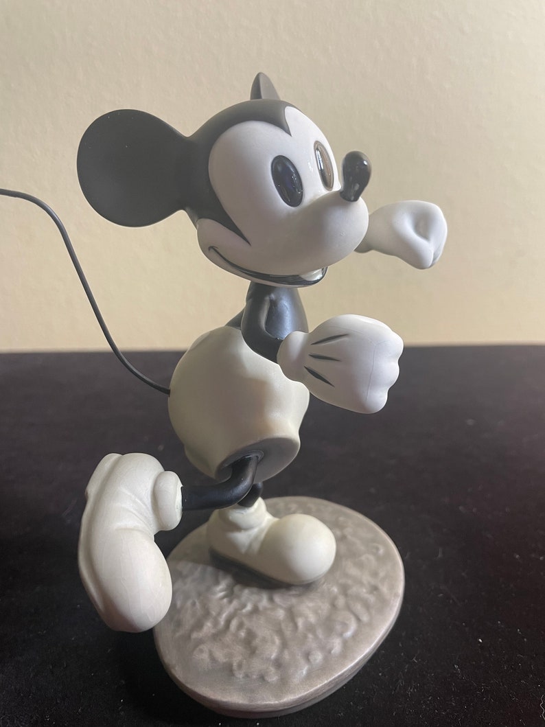 May include: A black and white ceramic figurine of Mickey Mouse running. The figurine is in a classic cartoon style and is posed with his right arm raised and his left leg forward. The figurine is standing on a gray base.