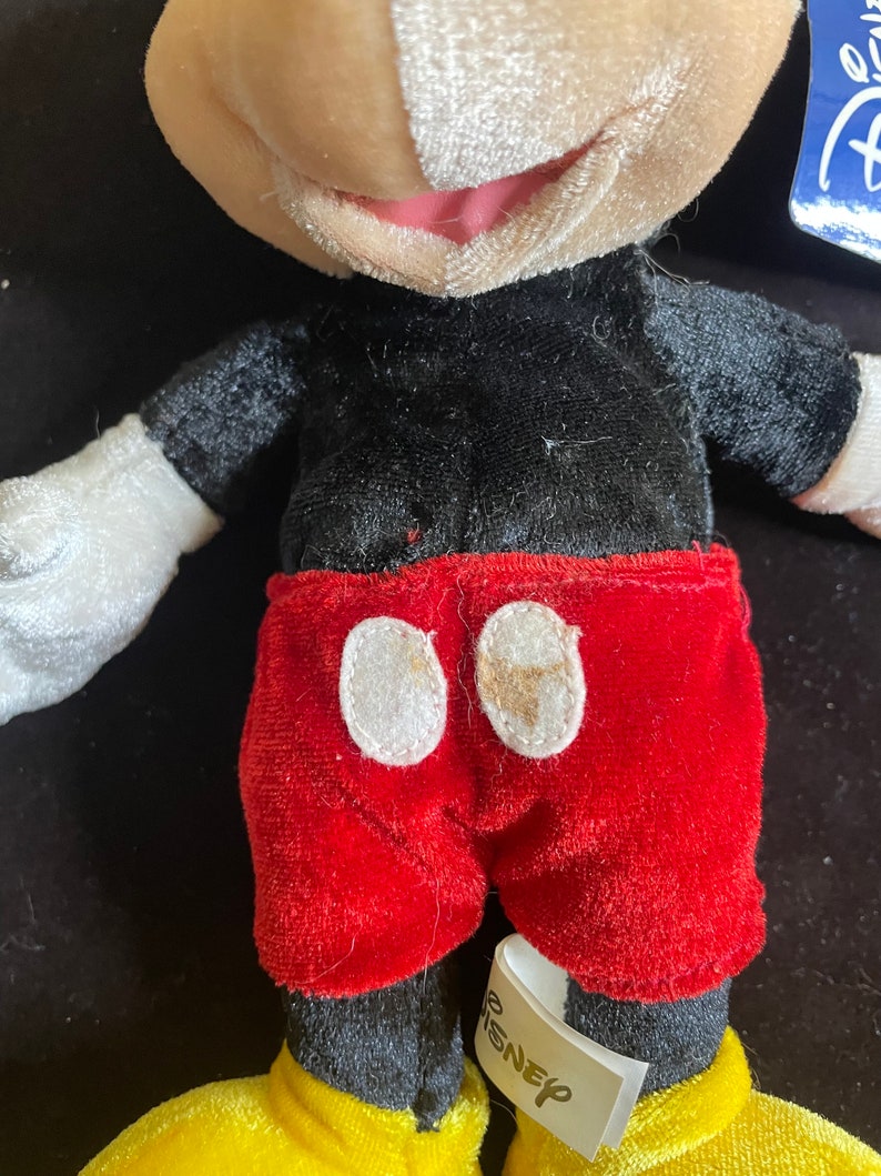 K&ouml;nnte beinhalten: Ein Pl&uuml;sch-Mickey-Mouse-Spielzeug mit schwarz-wei&szlig;em Fell, roten Shorts und gelben Schuhen. Das Spielzeug hat ein wei&szlig;es Etikett mit der Aufschrift "Disney".