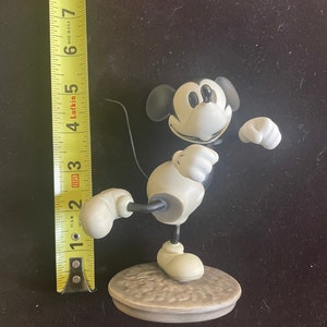 May include: A black and white ceramic figurine of Mickey Mouse in a classic pose. He is standing on a round base with a textured surface.
