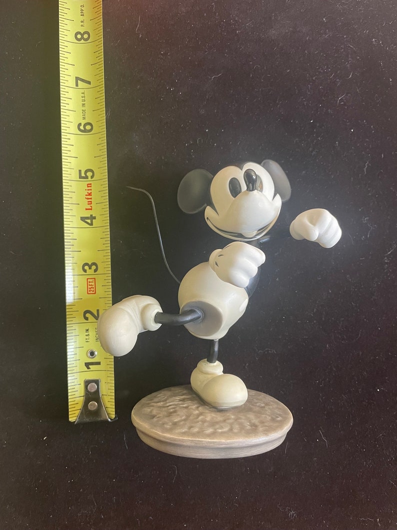 May include: A black and white ceramic figurine of Mickey Mouse in a classic pose. He is standing on a round base with a textured surface.