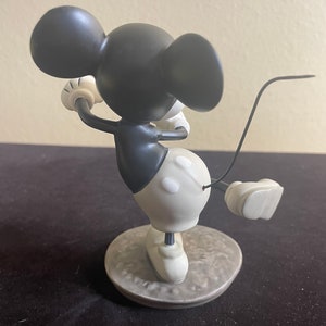 May include: A black and white ceramic figurine of Mickey Mouse playing golf. Mickey is wearing red shorts and white shoes. He is holding a golf club in his right hand and has a golf ball in his left hand. He is standing on a gray base.