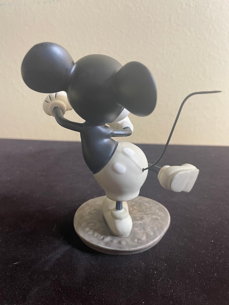 May include: A black and white ceramic figurine of Mickey Mouse playing golf. Mickey is wearing red shorts and white shoes. He is holding a golf club in his right hand and has a golf ball in his left hand. He is standing on a gray base.