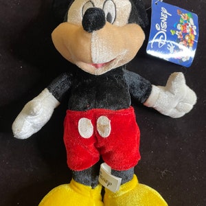 K&ouml;nnte beinhalten: Ein Pl&uuml;sch-Mickey-Mouse-Spielzeug mit schwarzem Fell, einem rot-wei&szlig;en Hemd und gelben Schuhen. Das Spielzeug hat ein Disney-Etikett am Arm.