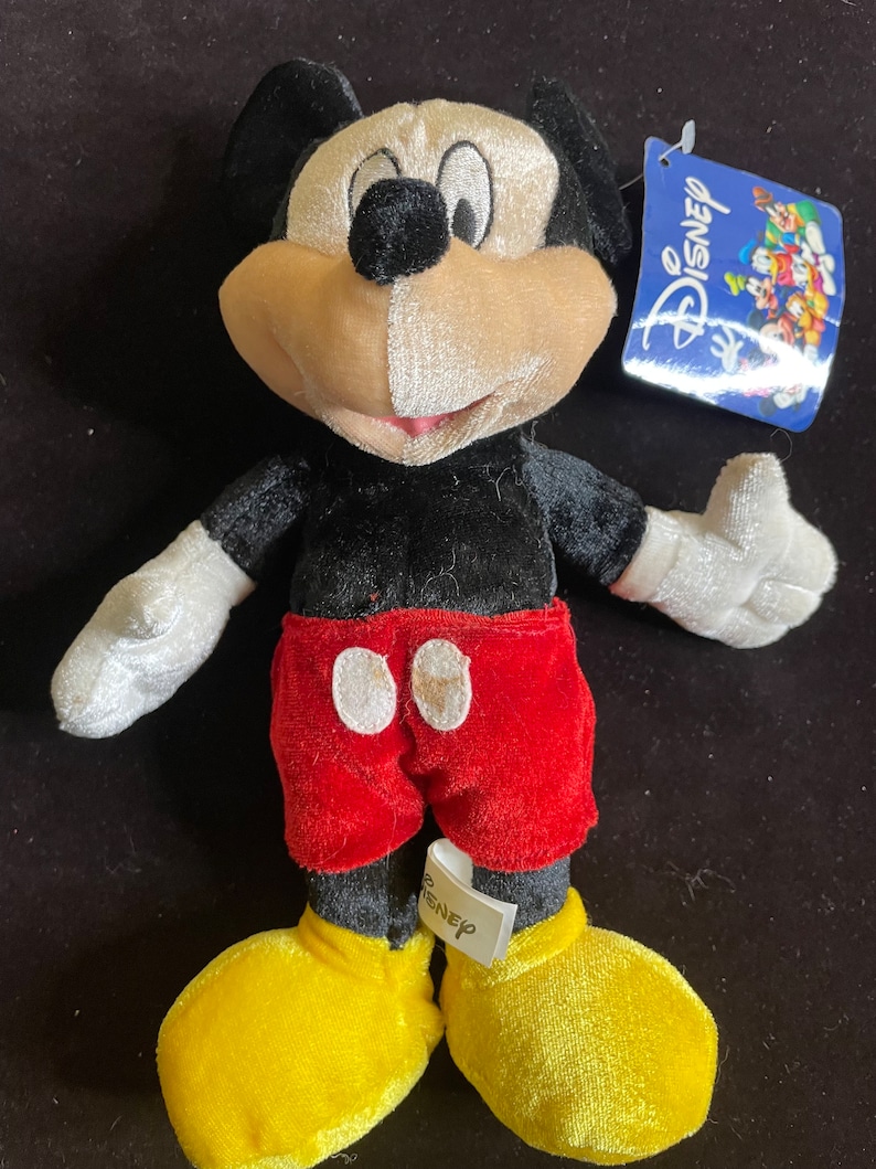 K&ouml;nnte beinhalten: Ein Pl&uuml;sch-Mickey-Mouse-Spielzeug mit schwarzem Fell, einem rot-wei&szlig;en Hemd und gelben Schuhen. Das Spielzeug hat ein Disney-Etikett am Arm.