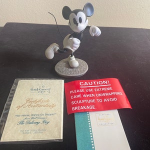 May include: A black and white figurine of Mickey Mouse in a running pose. The figurine is on a base with a certificate of authenticity and a warning label that reads "CAUTION! PLEASE USE EXTREME CARE WHEN UNWRAPPING SCULPTURE TO AVOID BREAKAGE."