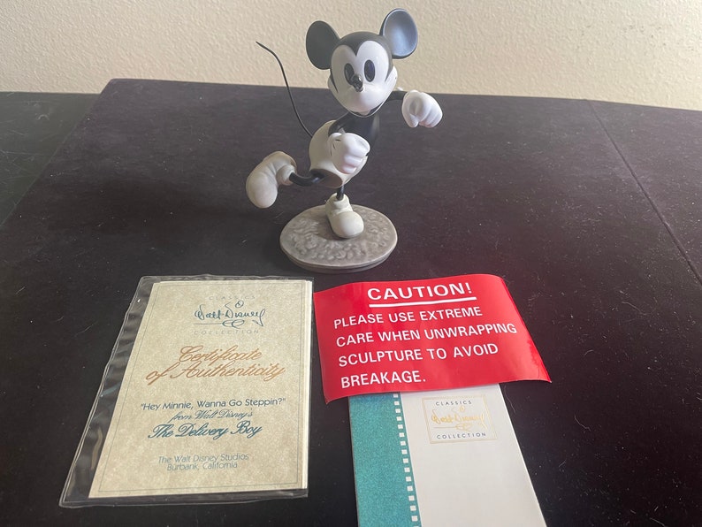 May include: A black and white figurine of Mickey Mouse in a running pose. The figurine is on a base with a certificate of authenticity and a warning label that reads "CAUTION! PLEASE USE EXTREME CARE WHEN UNWRAPPING SCULPTURE TO AVOID BREAKAGE."