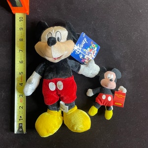 K&ouml;nnte beinhalten: Zwei Pl&uuml;sch-Mickey-Mouse-Spielzeuge. Der gr&ouml;&szlig;ere Mickey tr&auml;gt eine rote Hose mit wei&szlig;en Punkten und gelbe Schuhe. Der kleinere Mickey tr&auml;gt eine rote Hose mit wei&szlig;en Punkten und rote Schuhe. Beide Mickey-Mouse-Spielzeuge haben schwarze Ohren und schwarze Augen.