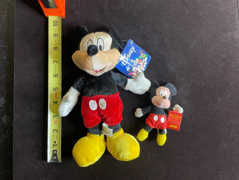 K&ouml;nnte beinhalten: Zwei Pl&uuml;sch-Mickey-Mouse-Spielzeuge. Der gr&ouml;&szlig;ere Mickey tr&auml;gt eine rote Hose mit wei&szlig;en Punkten und gelbe Schuhe. Der kleinere Mickey tr&auml;gt eine rote Hose mit wei&szlig;en Punkten und rote Schuhe. Beide Mickey-Mouse-Spielzeuge haben schwarze Ohren und schwarze Augen.