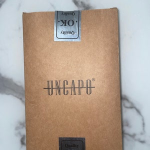 Könnte beinhalten: Ein brauner Karton mit dem Text "UNCAPO" in Schwarz gedruckt. Der Karton hat zwei silberne Aufkleber mit der Aufschrift "Quality OK. Quality".