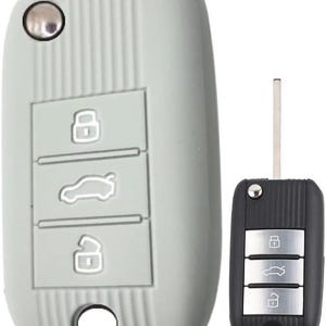 Könnte beinhalten: Eine hellgraue Autoschlüssel-Fernbedienung und ein schwarzer Autoschlüssel. Die graue Hülle hat drei Tasten mit Symbolen zum Ver- und Entriegeln sowie zum Öffnen des Kofferraums. Der schwarze Schlüssel hat die gleichen drei Tasten.