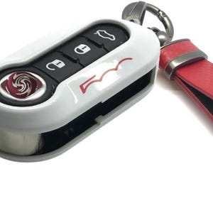 Könnte beinhalten: Weißer Autoschlüsselanhänger mit schwarzen Knöpfen und einem roten Akzent, befestigt an einem roten Lederband. Der Schlüsselanhänger hat einen silbernen Schlüssel und ein Logo mit rotem Wirbeldesign.