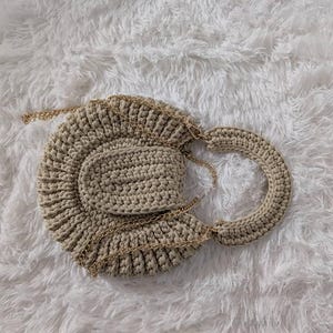 May include: A beige crocheted handbag with a circular design and a small matching pouch. The bag features a gold chain strap and a rounded handle. The bag is made of a textured yarn.