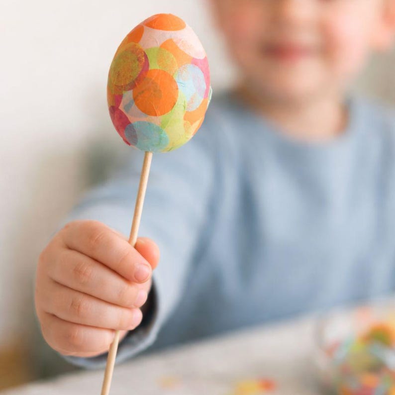 May include: A decorated Easter egg on a wooden stick. The egg is adorned with a collage of orange, pink, green, and blue circles. The egg is held by a person wearing a blue long-sleeve top.