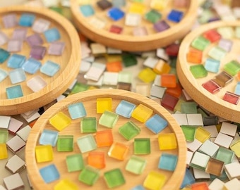May include: Four round wooden coasters with colourful mosaic tiles. The coasters are made of light-coloured wood and are filled with small, square tiles in shades of blue, green, yellow, orange, and purple. The tiles are scattered around the coasters.