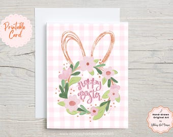Könnte beinhalten: Druckbare Karte mit einem rosa-weißen Gingham-Hintergrund. Ein rosafarbener Blumenkranz mit einem Kupfer-Hasenohren-Design umgibt den Text "Frohe Ostern".