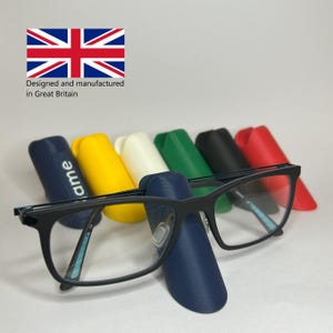 May include: Black framed eyeglasses with clear lenses are positioned in front of a row of colourful cases. The cases are in blue, yellow, white, green, black, and red. The text "Designed and manufactured in Great Britain" is visible, along with the Union Jack flag.