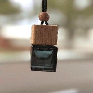 Puede incluir: Un ambientador para coche con un frasco de vidrio oscuro y una tapa de madera. El ambientador está suspendido por un cordón negro con una cuenta de madera. El fondo está borroso, sugiriendo un entorno al aire libre.