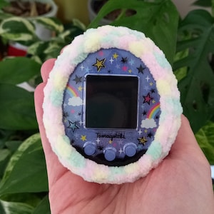 May include: A Tamagotchi digital pet with a blue face featuring stars, rainbows, and the Tamagotchi logo. It is encased in a fluffy, pastel-colored cover. The device has a black screen and three gray buttons.