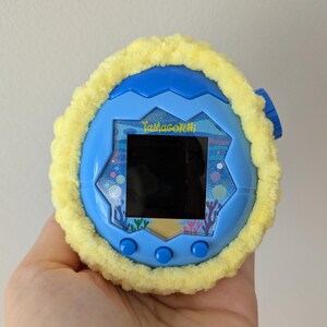 May include: A blue Tamagotchi virtual pet with a yellow fuzzy cover. The device has a black screen and three blue buttons. The Tamagotchi is held in a hand against a neutral background.