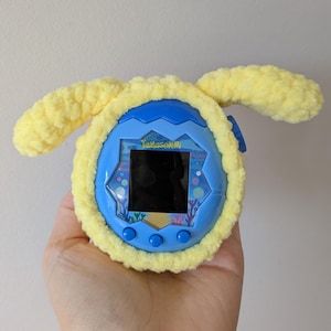 May include: A blue Tamagotchi digital pet with a yellow plush cover featuring two ears. The screen displays a black screen. The text "Tamagotchi" is visible on the screen. The device is held in a hand.