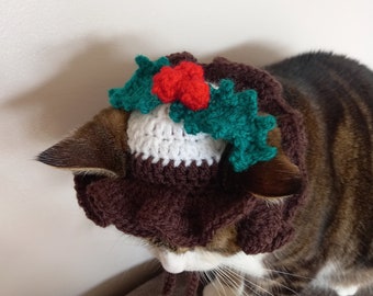 May include: A brown and white crocheted cat hat with a green holly wreath and red berries. The hat is worn by a tabby cat.
