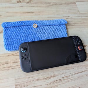 May include: A blue, crocheted case with a wooden button closure next to a black handheld gaming console. The case is made of a soft, textured yarn. The console has black controls with red and blue accents.