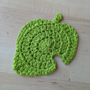 May include: A green crocheted leaf-shaped coaster. The coaster is made of yarn and has a textured surface.