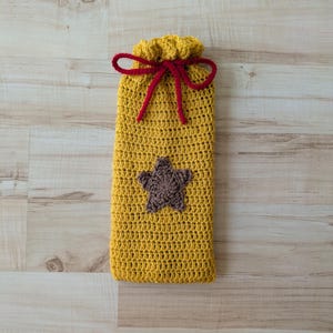 May include: A handmade, yellow crochet bag with a brown star applique. The bag has a drawstring closure with a red bow. The bag is made of a soft, textured yarn.