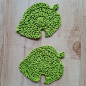 May include: Two green crocheted leaf-shaped coasters. The coasters are made with a simple stitch pattern and have a rounded shape.