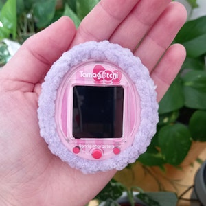 May include: A pink and purple Tamagotchi with a fluffy purple cover. The screen displays the text "Tamagotchi" and "Sanrio characters".