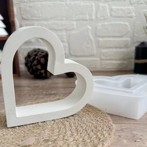 Könnte beinhalten: Zwei weiße herzförmige Formen, eine größer als die andere, werden auf einer braunen Oberfläche gezeigt. Die größere Form besteht aus einem strukturierten Material, während die kleinere Form aus einem glatten, flexiblen Material besteht.