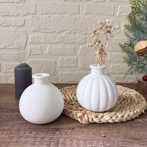 Könnte beinhalten: Zwei weiße Keramikvasen. Eine ist rund, die andere hat eine gerippte Kürbisform. Die Kürbisvase enthält Trockenblumen. Links steht eine dunkelgraue Kerze. Die Vasen stehen auf einer gewebten Matte.