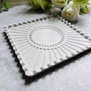Peut inclure: Un plateau carré blanc avec un bord festonné et une découpe circulaire au centre. Le plateau a un motif en forme de soleil rayonnant du centre.