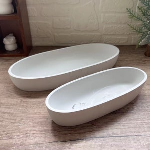 Op de afbeelding: Twee witte ovale keramische schalen, een groter dan de andere, op een houten oppervlak. De kleinere schaal heeft een reliëfdesign aan de binnenkant.