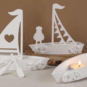 Puede incluir: Candelabros decorativos blancos con forma de barco. Uno tiene un corazón recortado en la vela, otro una figura de pájaro y un tercero sostiene una vela encendida. Los barcos tienen elementos decorativos.