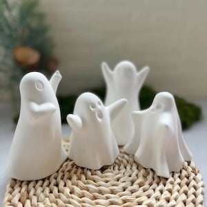 Può includere: Quattro figurine di fantasmi in ceramica bianca con pose diverse, sedute su una superficie di vimini intrecciato.