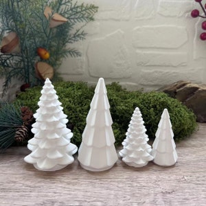 以下が含まれることがあります： さまざまなサイズの白いセラミック製のクリスマスツリーのオーナメントが4つ。木は、木製の表面に、大きい順に並んでいます。