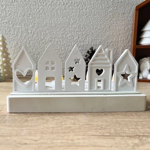Może przedstawiać: Biały drewniany świecznik w kształcie rzędu domków z wyciętymi sercami i gwiazdami. Świecznik ma podstawę i jest przeznaczony do trzymania świeczek typu tealight.
