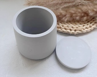 Puede incluir: Un recipiente cilíndrico gris claro con una tapa a juego. El recipiente está hecho de un material liso y mate y tiene un diseño simple y moderno.