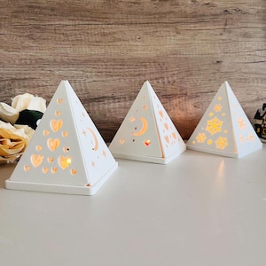 Può includere: Tre portacandele bianchi a forma di piramide con diversi disegni ritagliati. Il primo portacandele ha una mezzaluna e stelle, il secondo ha cuori e il terzo ha fiocchi di neve. Ogni portacandele ha una piccola candela all'interno, che proietta un bagliore caldo.