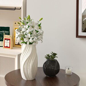 May include: A white ceramic vase with a textured, wave-like design holds a bouquet of white lilies. A small black decorative pot with a plant and a lit candle in a glass holder sit on a round, dark brown table.
